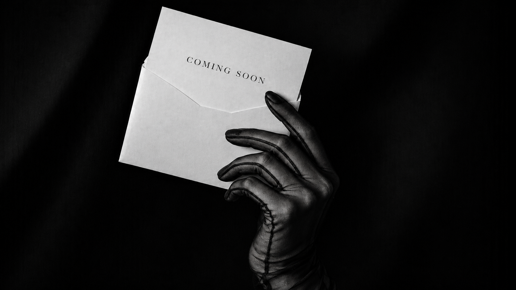 A sheer black-gloved hand holding an elegant white envelope with a card that says Coming Soon.